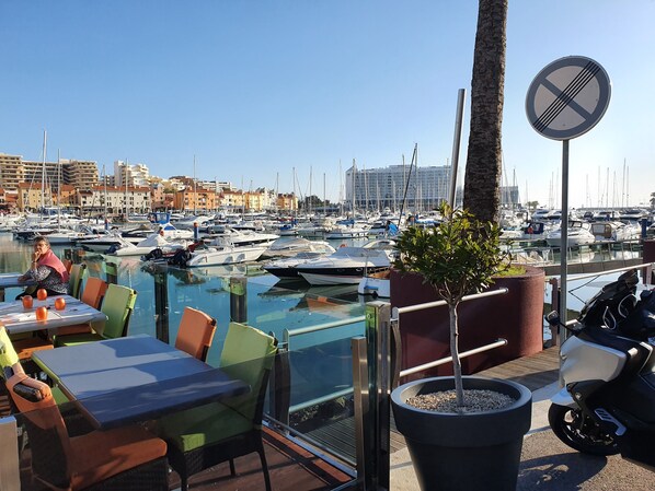Uma marina com barcos, uma área de refeições com mesas e cadeiras, e uma placa de proibido estacionar.