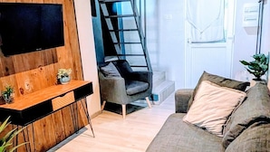 A modern living room with a wooden accent wall, a flat-screen TV, a dark grey sofa, a staircase, and a small table with plants. - Appartement Lila (Carcassonne)
