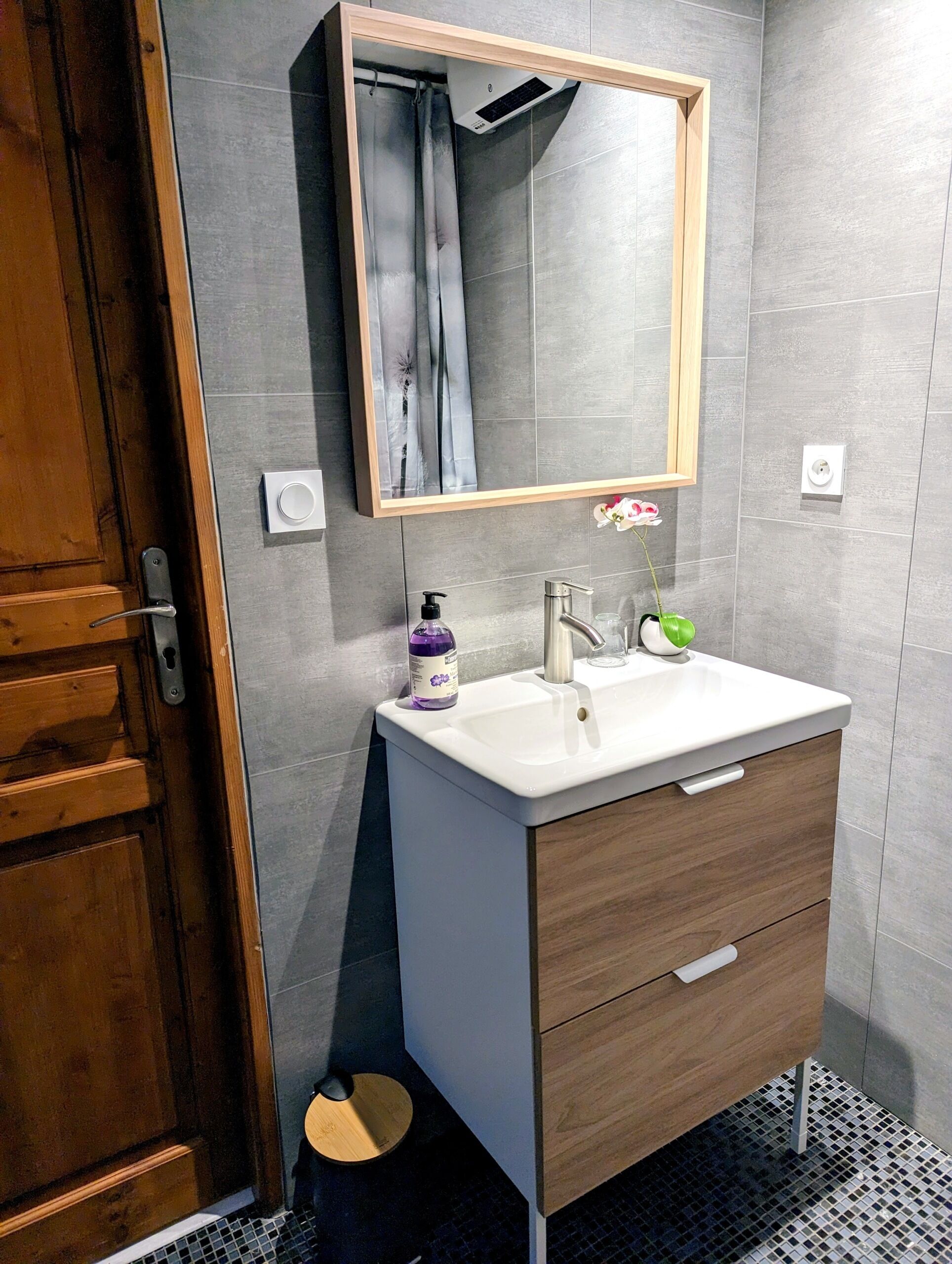 Une salle de bain avec une porte en bois, un lavabo blanc, un miroir et un petit tabouret rond.
