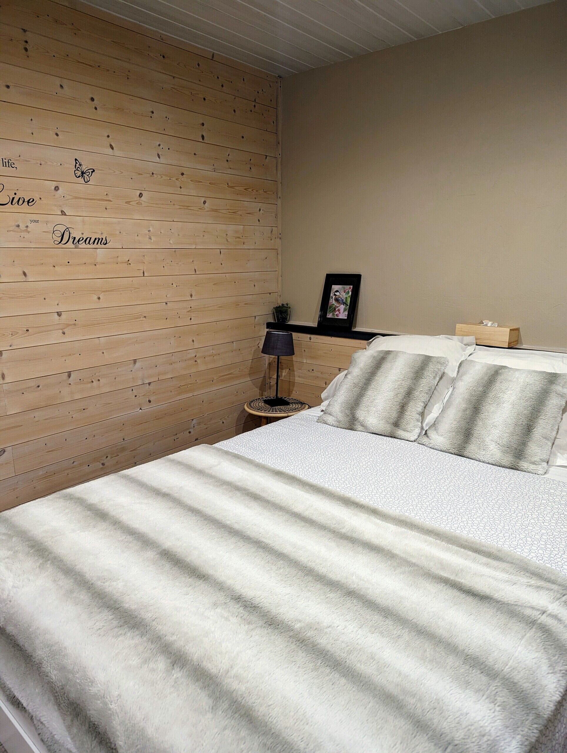 A bedroom with a wooden wall, a bed with a patterned bedspread, a bedside lamp, and a small shelf with a picture frame and a box.