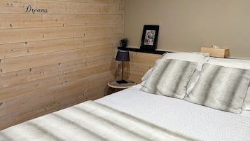 A bedroom with a wooden wall, a bed with a patterned bedspread, a bedside lamp, and a small shelf with a picture frame and a box.
