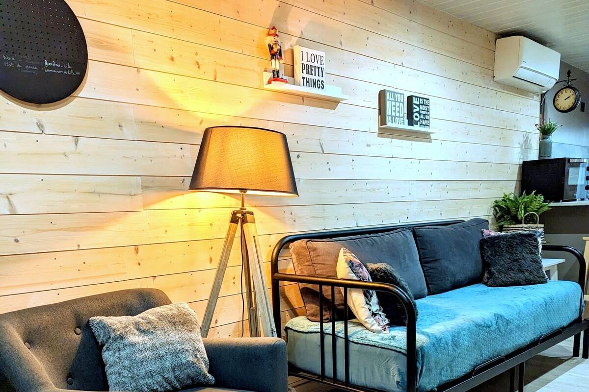 A cozy living room with a dark blue armchair, a blue sofa, a floor lamp, and a wooden wall with decorative items.