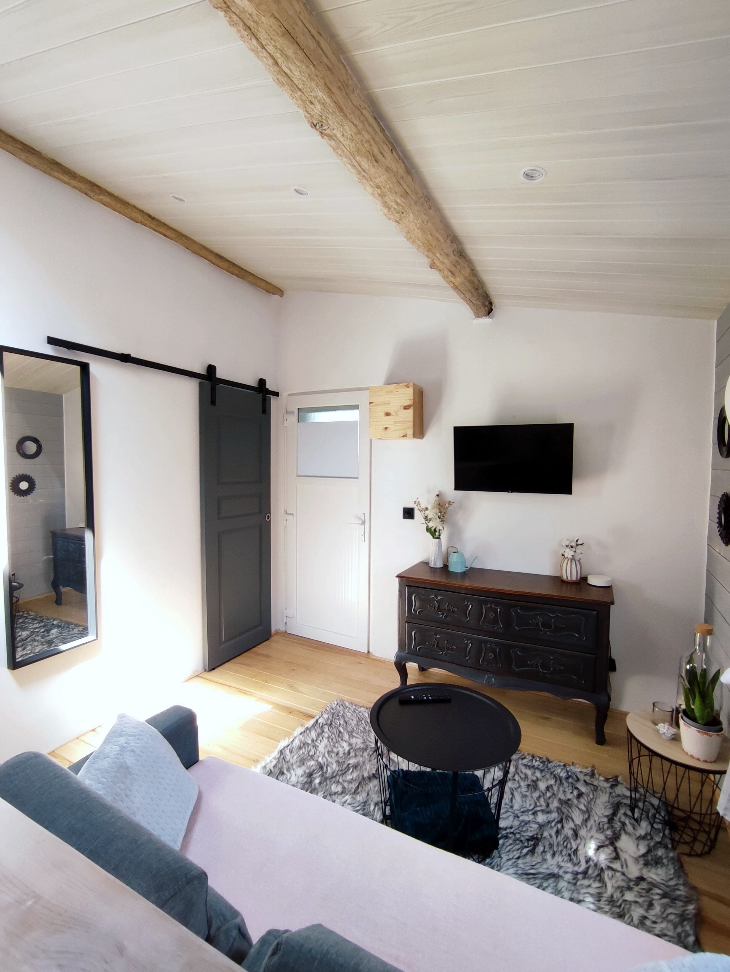 A living room with a sofa, a coffee table, a TV mounted on the wall, a wooden cabinet, and a door leading to another room.