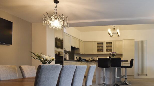 Un comedor moderno con una mesa de madera y sillas grises tapizadas, una araña y una cocina al fondo.