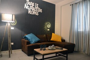 A living room with a brown sofa, blue and yellow cushions, a coffee table with glasses and a bowl, a floor lamp, and a wall with a motivational quote.