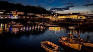 Uma vila à beira-rio ao entardecer, com barcos atracados, edifícios iluminados e um rio calmo refletindo as luzes.
