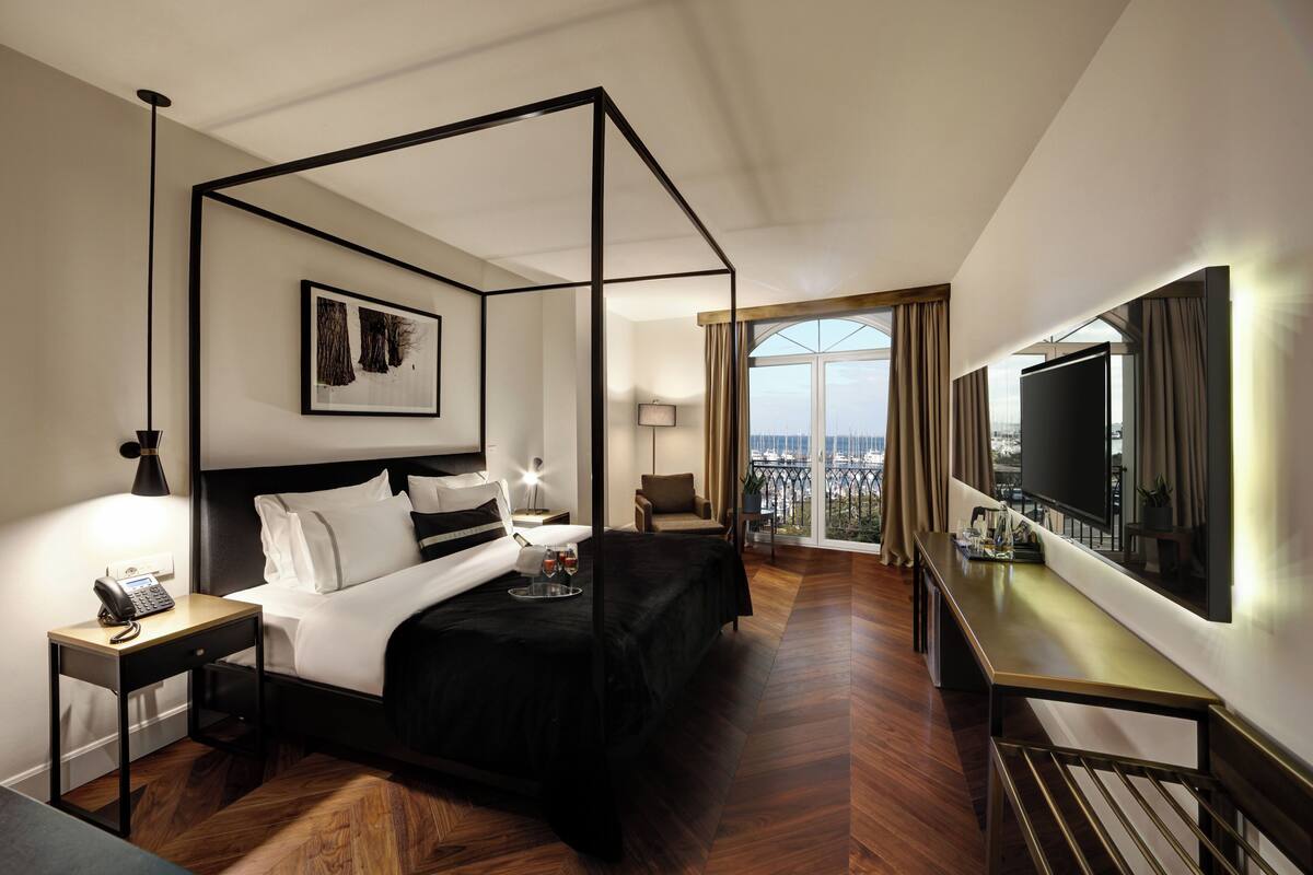 A modern hotel room with a four-poster bed, a flat-screen TV, a balcony with a city view, and a desk with a lamp.