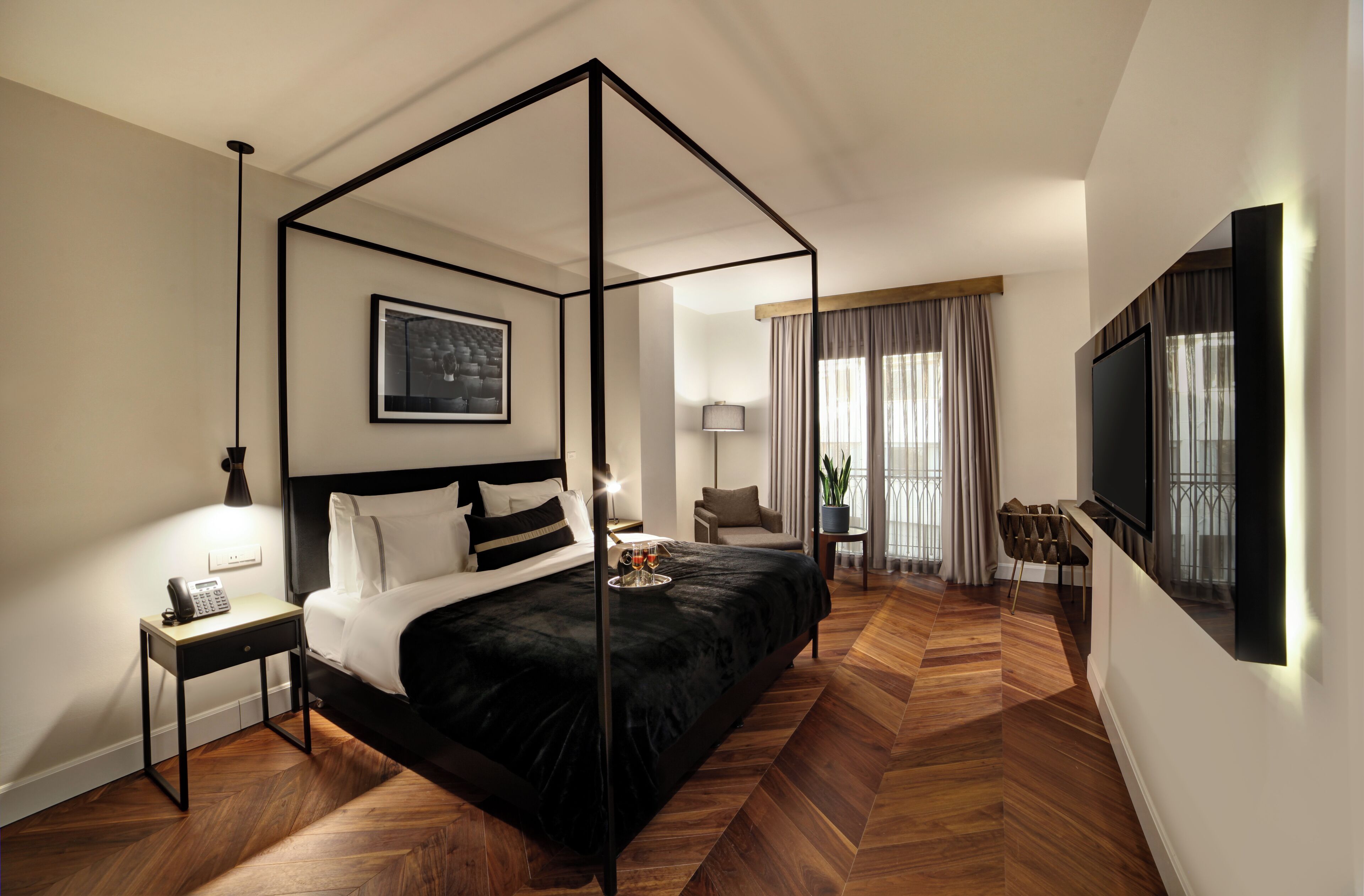 A modern hotel room with a four-poster bed, a flat-screen TV, a bedside table with a lamp, and a small table with a tray.