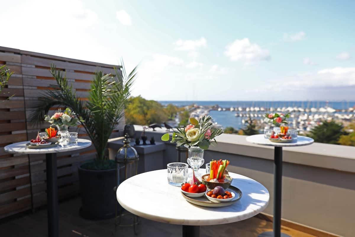 A terrace with round tables set for dining, featuring fresh fruits and flowers, overlooking a marina and cityscape.