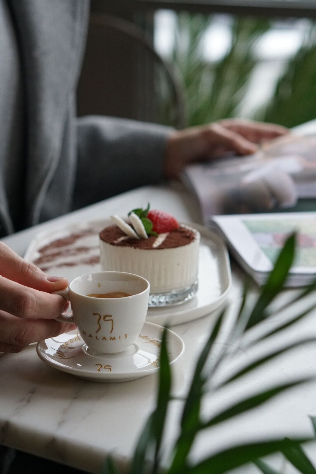 Une personne tenant une tasse portant le numéro 39, assise à une table devant un dessert et une assiette.