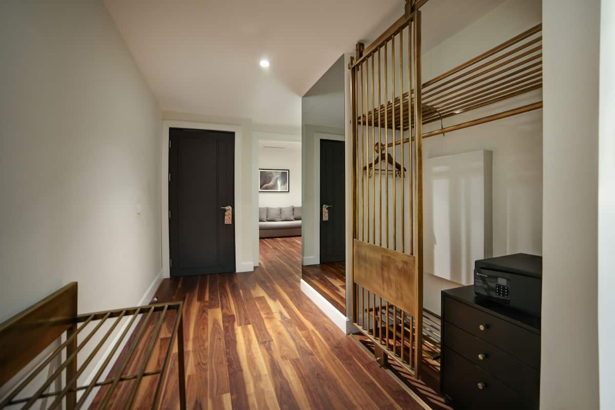 A hallway with a wooden floor, a black bench, a black dresser with a printer, and a sliding door leading to a room with a bed and a framed picture on the wall.