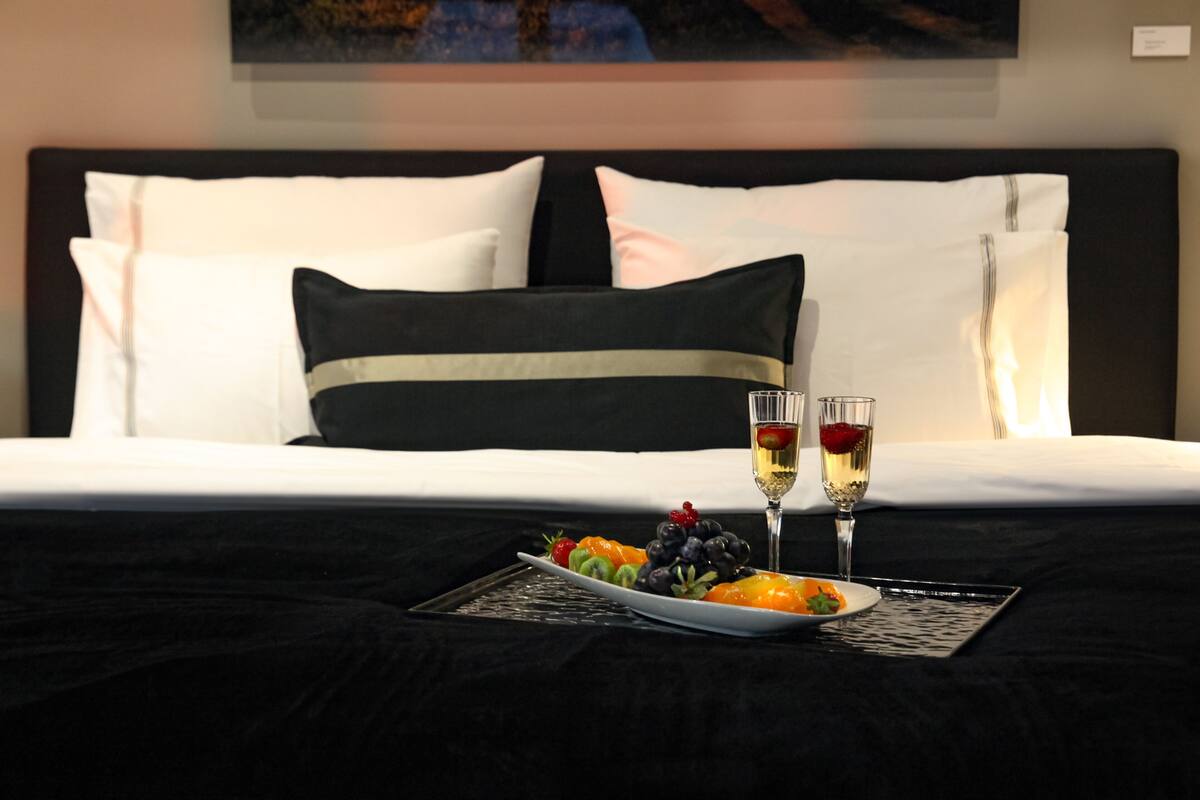 A neatly made bed with a black and white pillow arrangement, a fruit platter, and two glasses of wine on a tray.