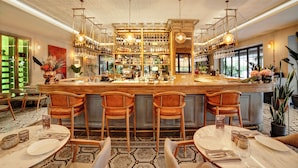Uma área de bar com balcão de madeira, vários banquetas e uma variedade de garrafas e copos em prateleiras.