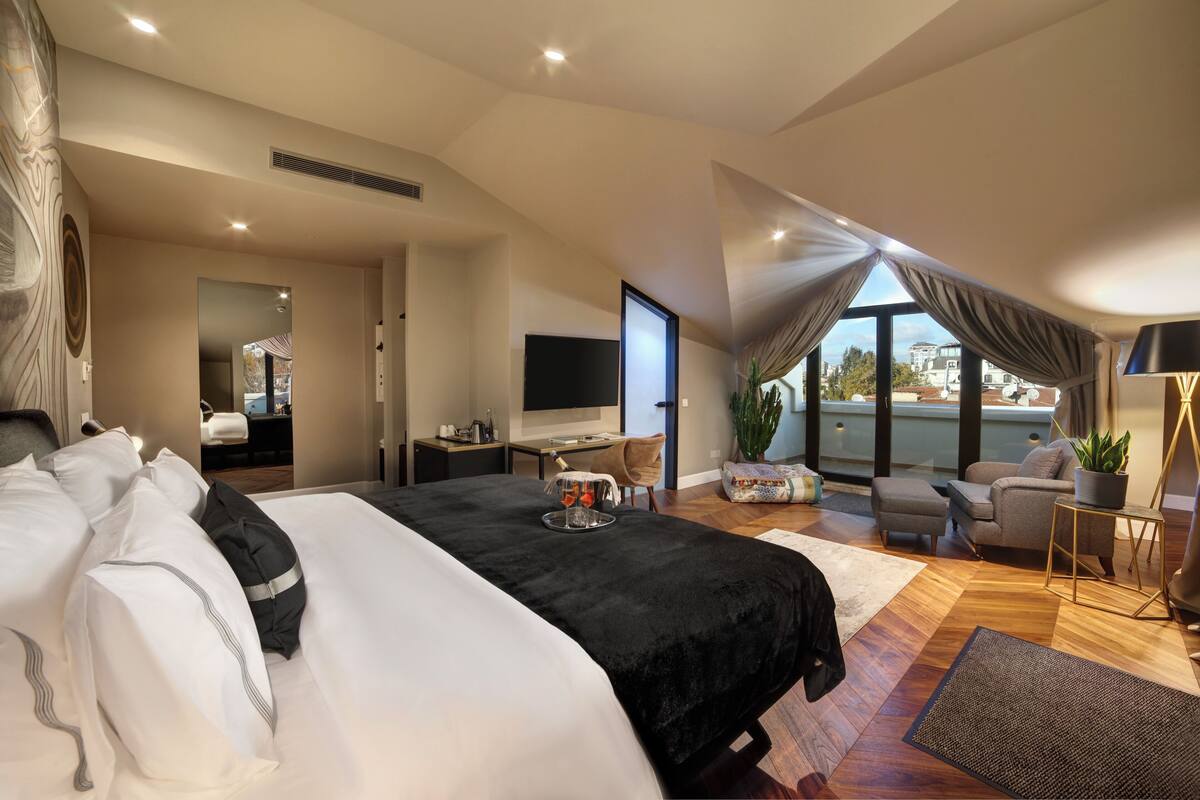 A spacious bedroom with a large bed, a desk, a TV, and a balcony with a view.