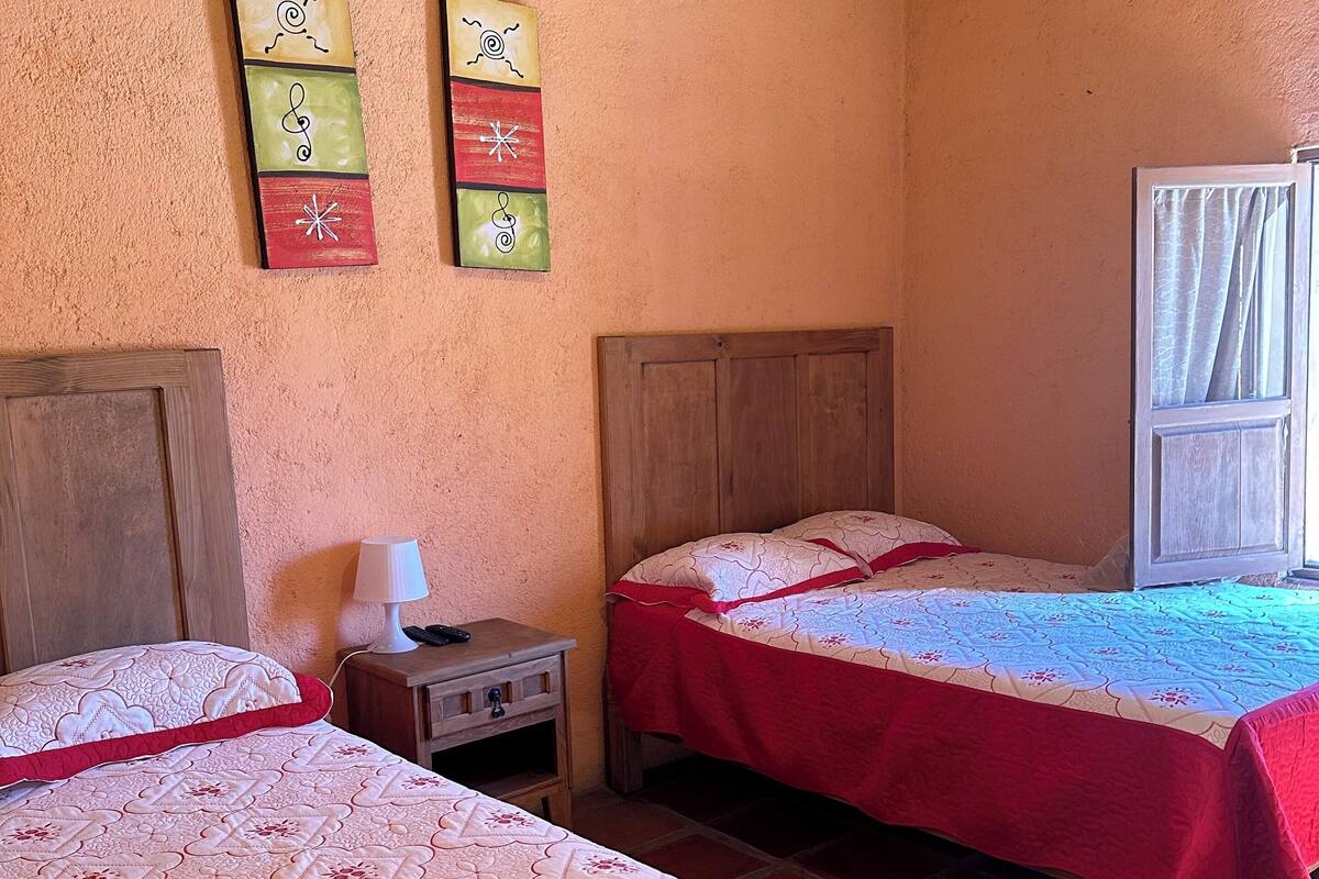 A room with two beds, wooden headboards, a nightstand with a lamp, and a window with curtains.
