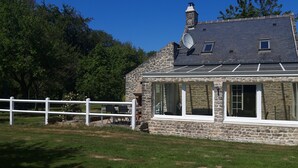 A stone house with a sloped roof, a satellite dish, and a white fence. - Charming cottage with a veranda facing south overlooking an orchard. (Colleville-sur-Mer)