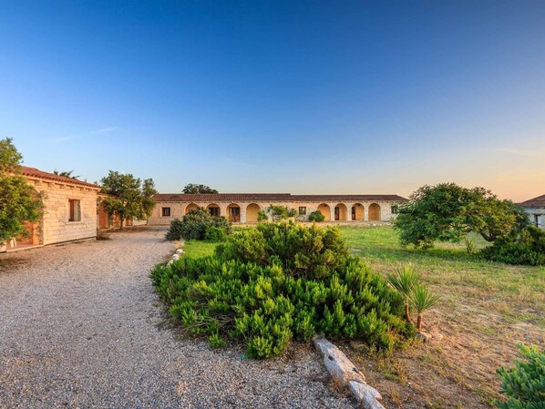 A courtyard with a building featuring a tiled roof and arched windows, surrounded by gravel paths and greenery.