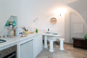 A compact kitchen with white cabinetry, a countertop with various kitchen items, a small dining table, and stools.