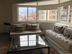 Ein modernes Wohnzimmer mit einer Couch, einem Glastisch und Blick auf eine Stadtkulisse durch große Fenster.