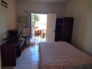 Une chambre avec un lit, un bureau équipé d’un ventilateur, une armoire et une vue sur un espace extérieur comprenant une porte et une table.