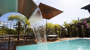 Ein Pool mit Wasserspiel, umgeben von einer Holzterrasse, Stühlen und Sonnenschirmen.