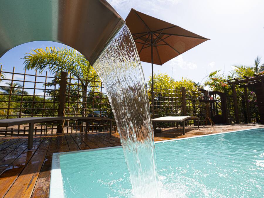 A pool with a water feature, surrounded by wooden deck, chairs, and umbrellas.