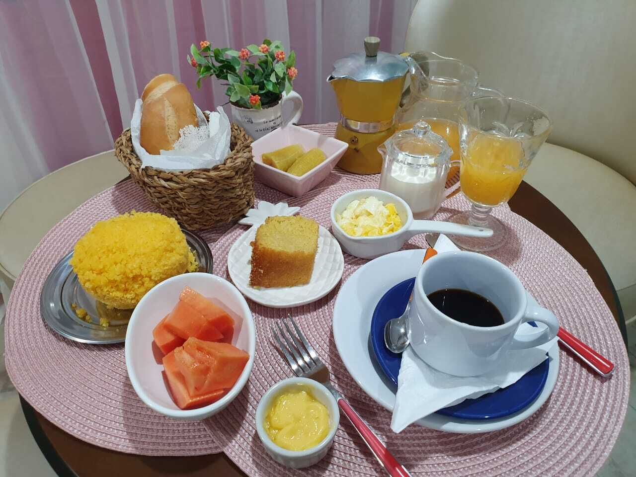 Et morgenbord fyldt med forskelligt mad, blandt andet brød, majs, kage og frugt, samt en kaffekop og glas med appelsinjuice.