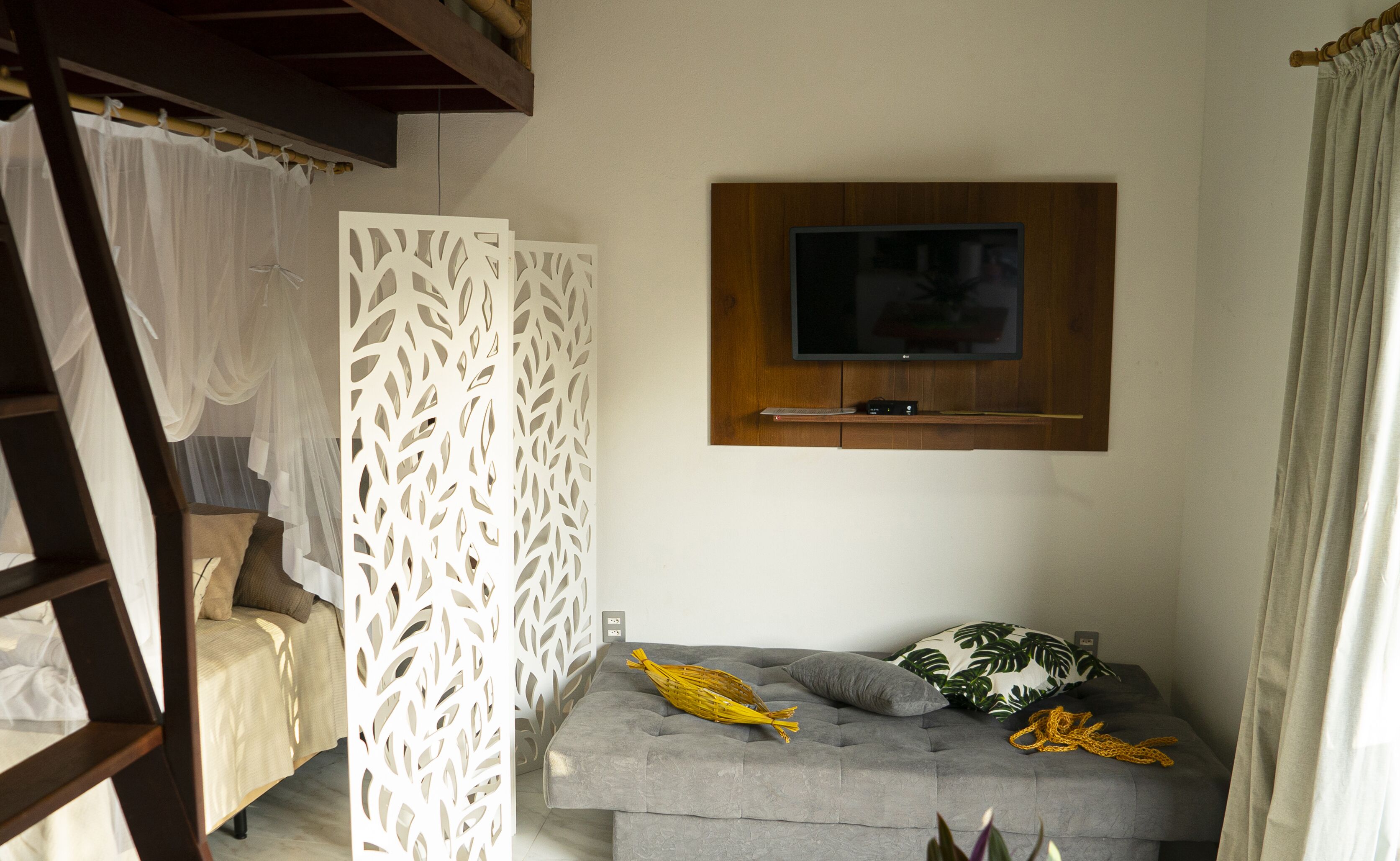 Quarto com sofá cinzento, televisão fixada em painel de madeira e divisória decorativa branca.
