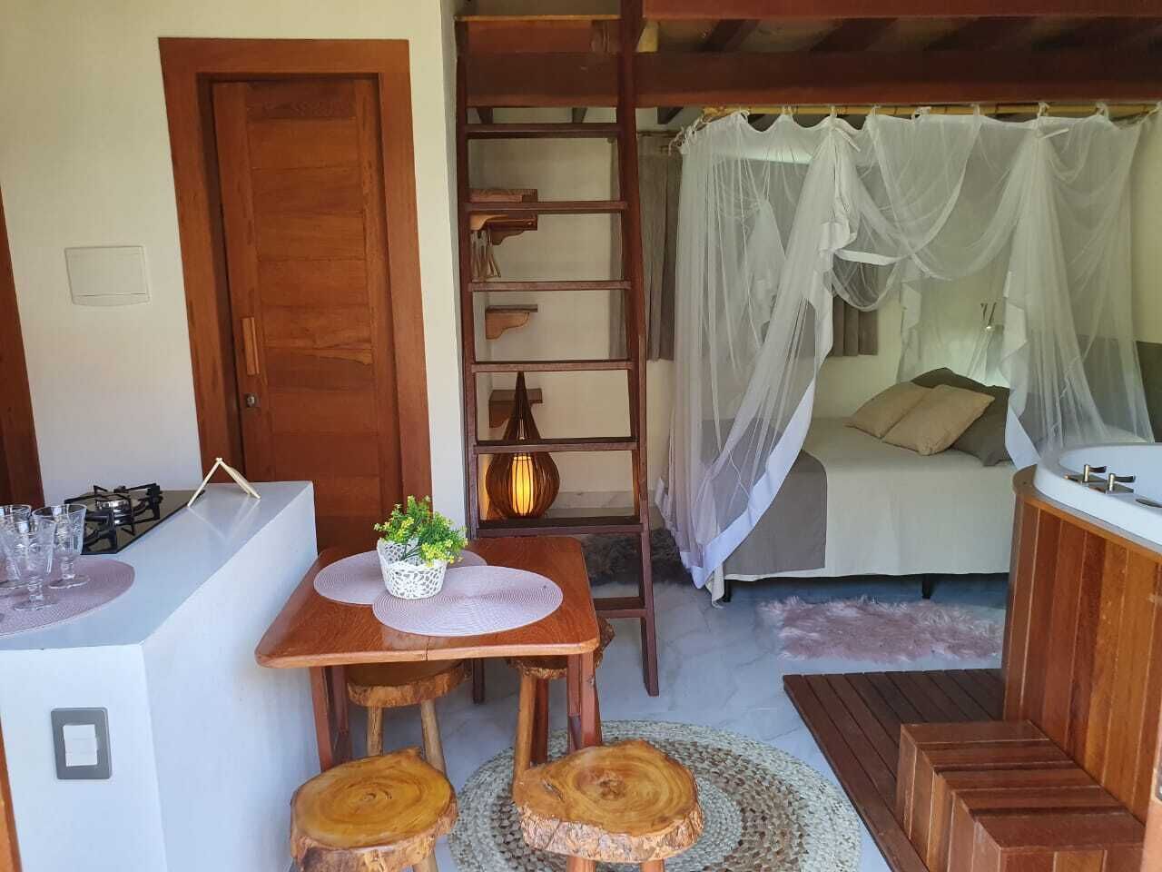 A cozy room with a bed under a canopy, a small table with chairs, a wooden ladder, and a kitchenette area.