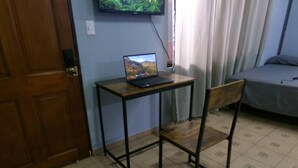 Une pièce avec un bureau en bois, un ordinateur portable, une chaise, un lit et un téléviseur fixé au mur.