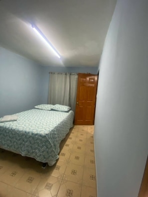 A small, single-bed room with a patterned bedspread, a wooden door, and a tiled floor.