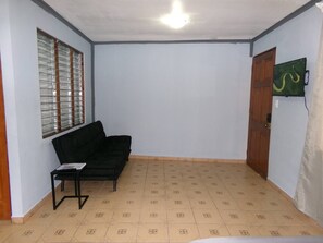 Une pièce avec un canapé noir, une petite table, une fenêtre avec des stores, une porte et un téléviseur fixé au mur.