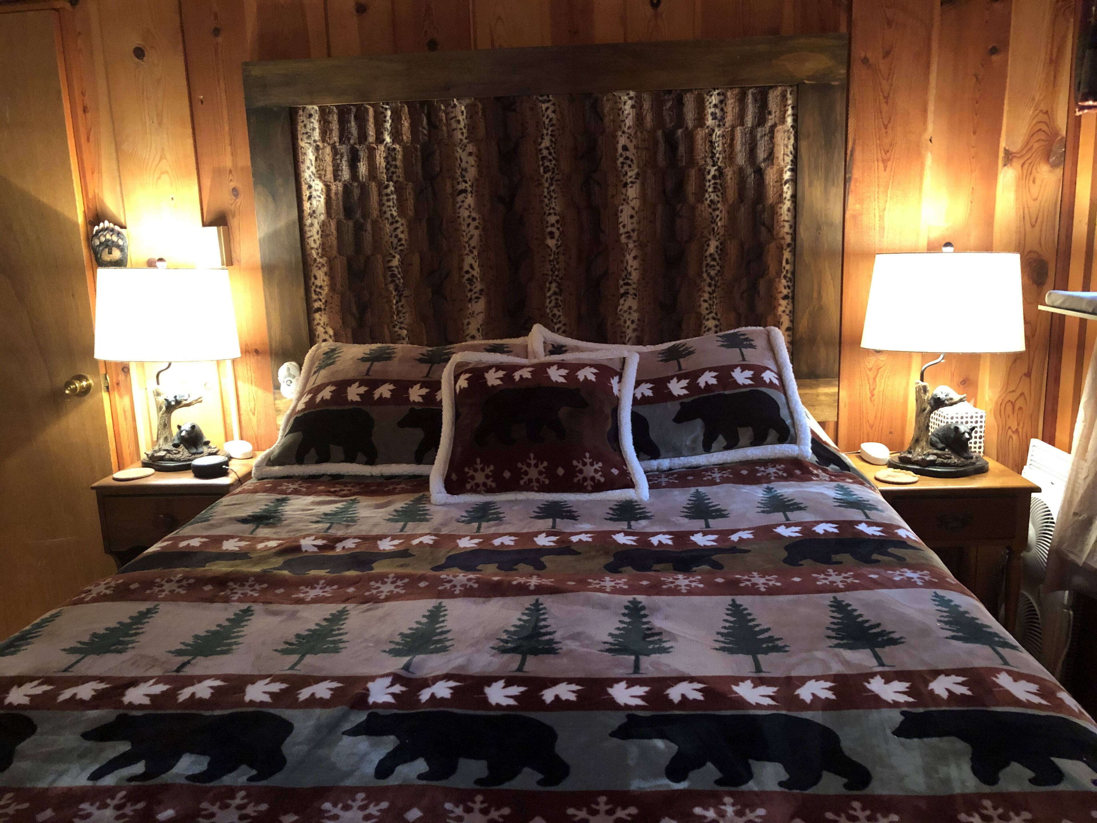 A cozy room with wooden paneling, a bed with a bear-themed blanket, two bedside lamps, and a nightstand with a lamp and a decorative item.