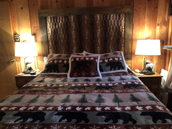 A cozy room with wooden paneling, a bed with a bear-themed blanket, two bedside lamps, and a nightstand with a lamp and a decorative item.