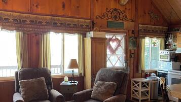 A cozy cabin interior with wooden walls, a chandelier, two armchairs, a dining table, and a kitchen area.