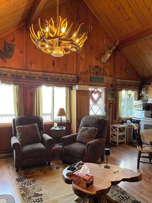 A cozy cabin interior with wooden walls, a chandelier, two armchairs, a dining table, and a kitchen area.