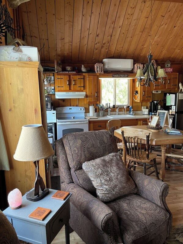 A cozy cabin interior with a wooden dining table, a comfortable armchair, and a kitchen area with wooden cabinets and a stove. - Black Bear Cozy Chalet, Lakeview, kayaks available for use (Lake Harmony)
