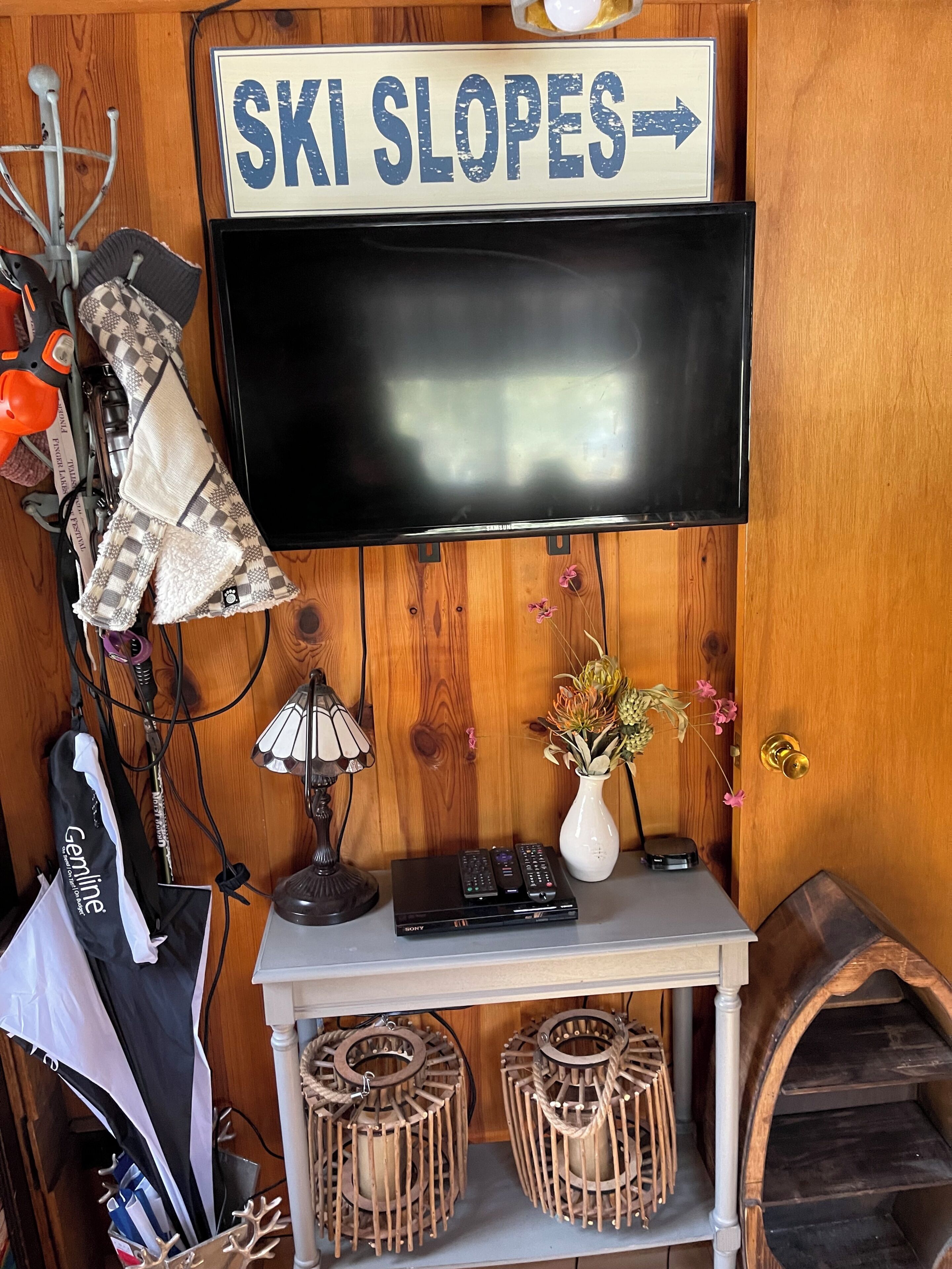 A room with a mounted flat-screen TV, a wooden wall, a sign reading 'SKI SLOPES,' a lamp, a vase with flowers, and a shelf with remote controls and a bag.