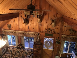 A cabin interior with a wooden ceiling, a ceiling fan, mounted animal heads, a quilted bed, and a window with a view of the outside.
