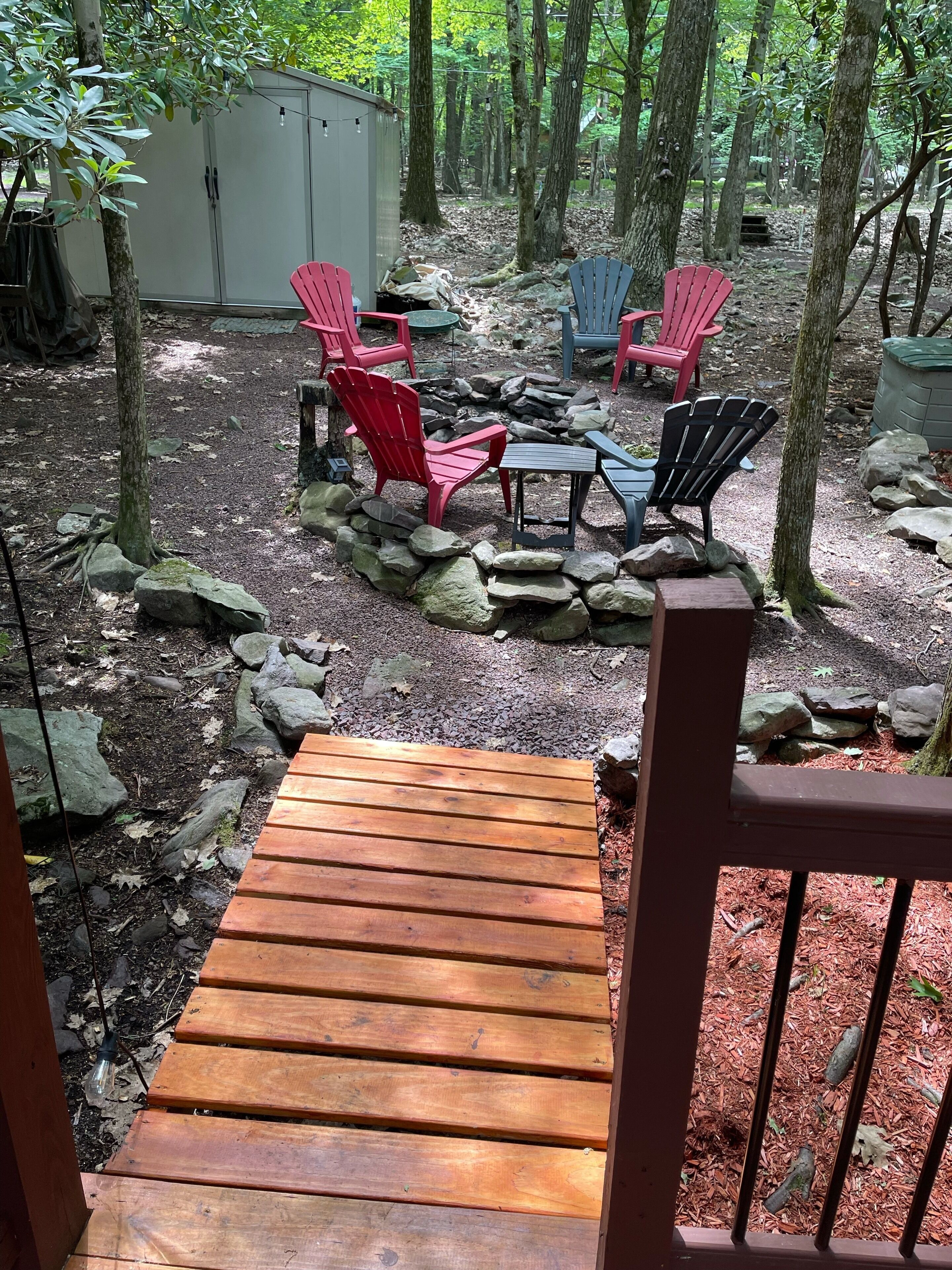 A wooden deck leading to a seating area with Adirondack chairs and a small table, surrounded by rocks and trees.