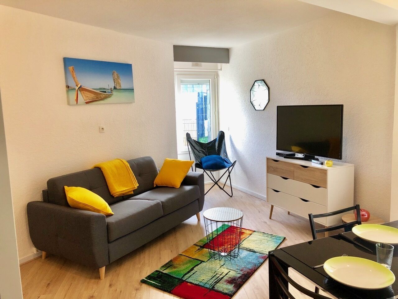 Uma sala de estar moderna com um sofá cinzento, almofadas amarelas, um tapete colorido, uma mesa de jantar com pratos, uma TV em um armário branco e um quadro de um barco na parede.