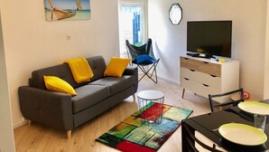A modern living room with a grey sofa, yellow cushions, a colorful rug, a dining table with plates, a TV on a white cabinet, and a painting of a boat on the wall. - Cosy cottage with terrace and private parking (Guidel)
