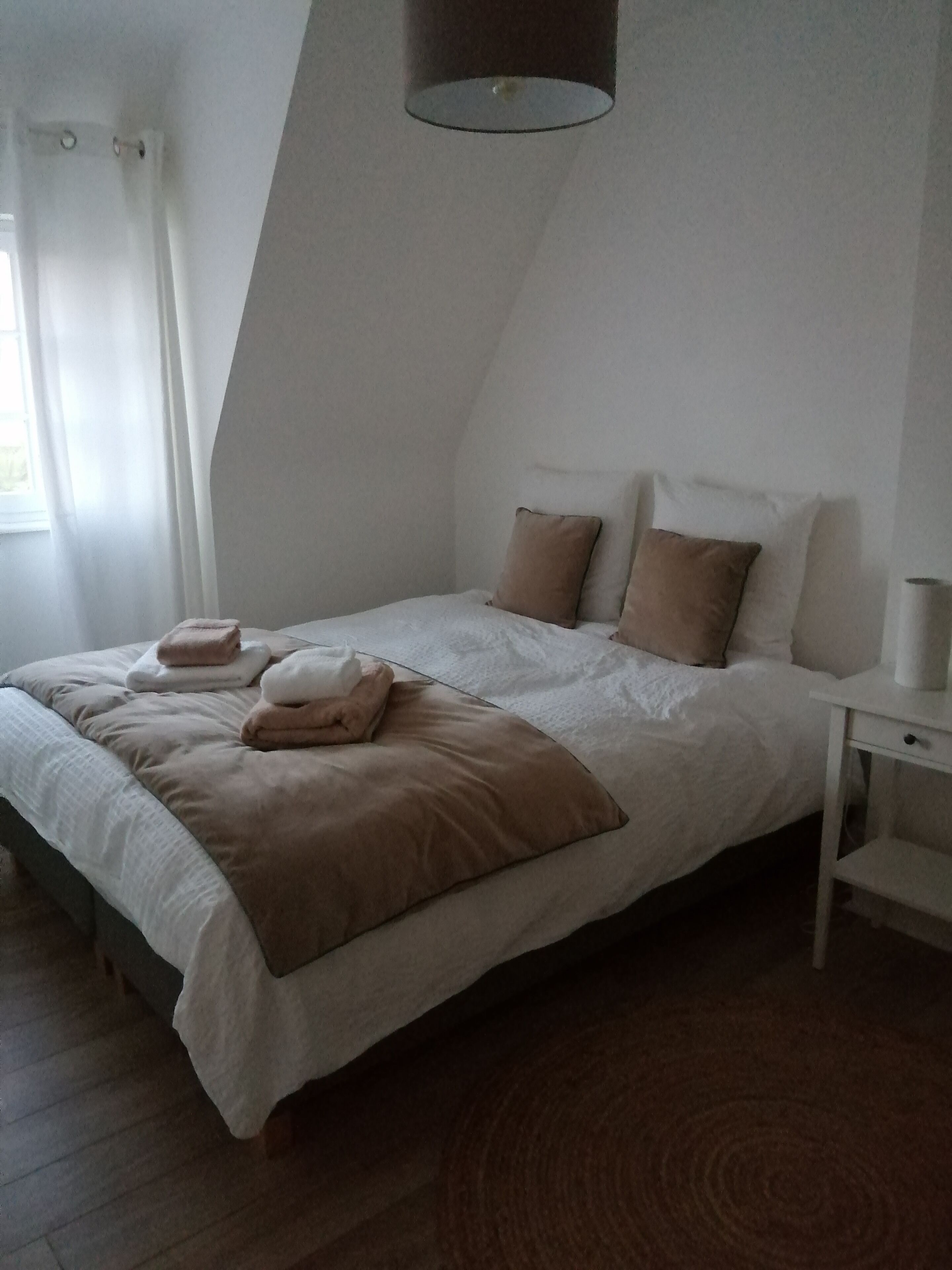 Een slaapkamer met een bed, kussens, een nachtkastje en een raam met gordijnen.
