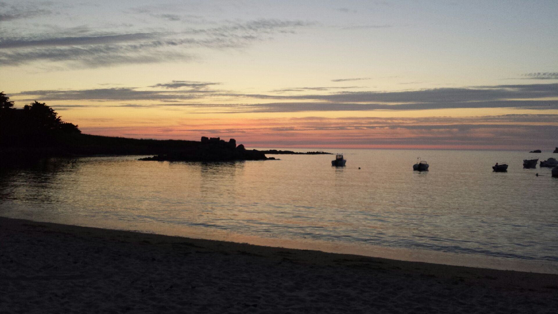 A calm beach with boats on the water and a sunset in the background.