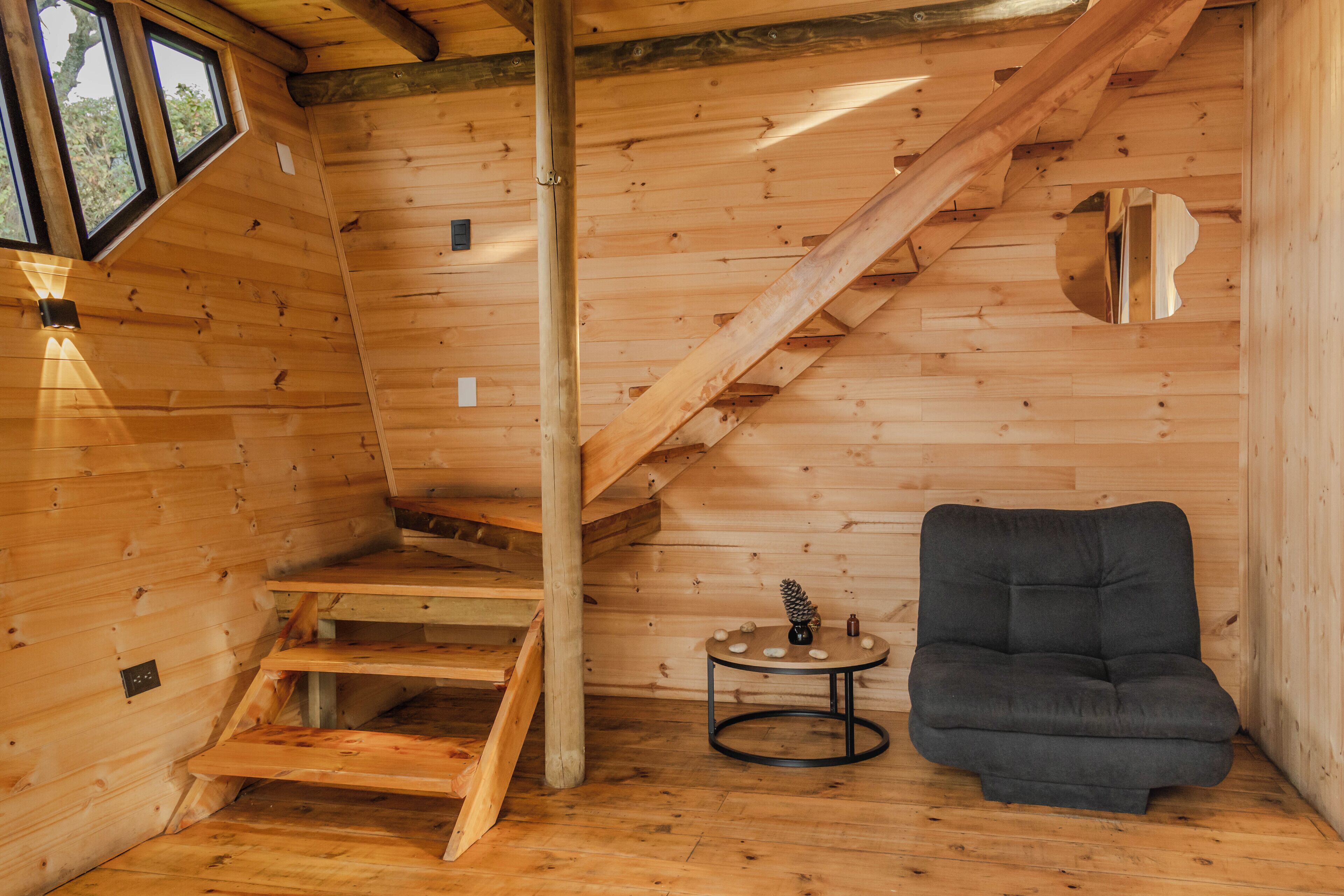 A wooden cabin interior with a staircase, a small round table with a decorative item, and a dark-colored sofa.