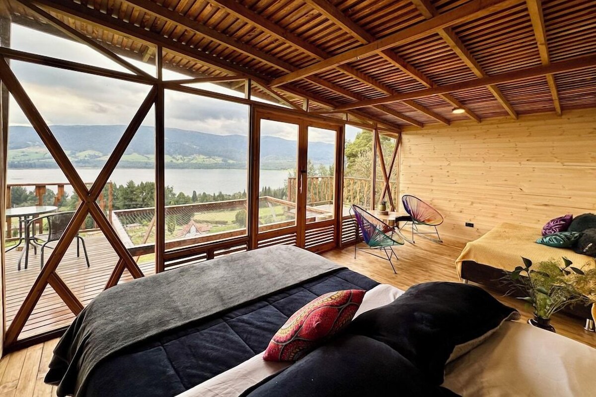 A room with a bed, a table, and chairs, large windows with a view of the outdoors.