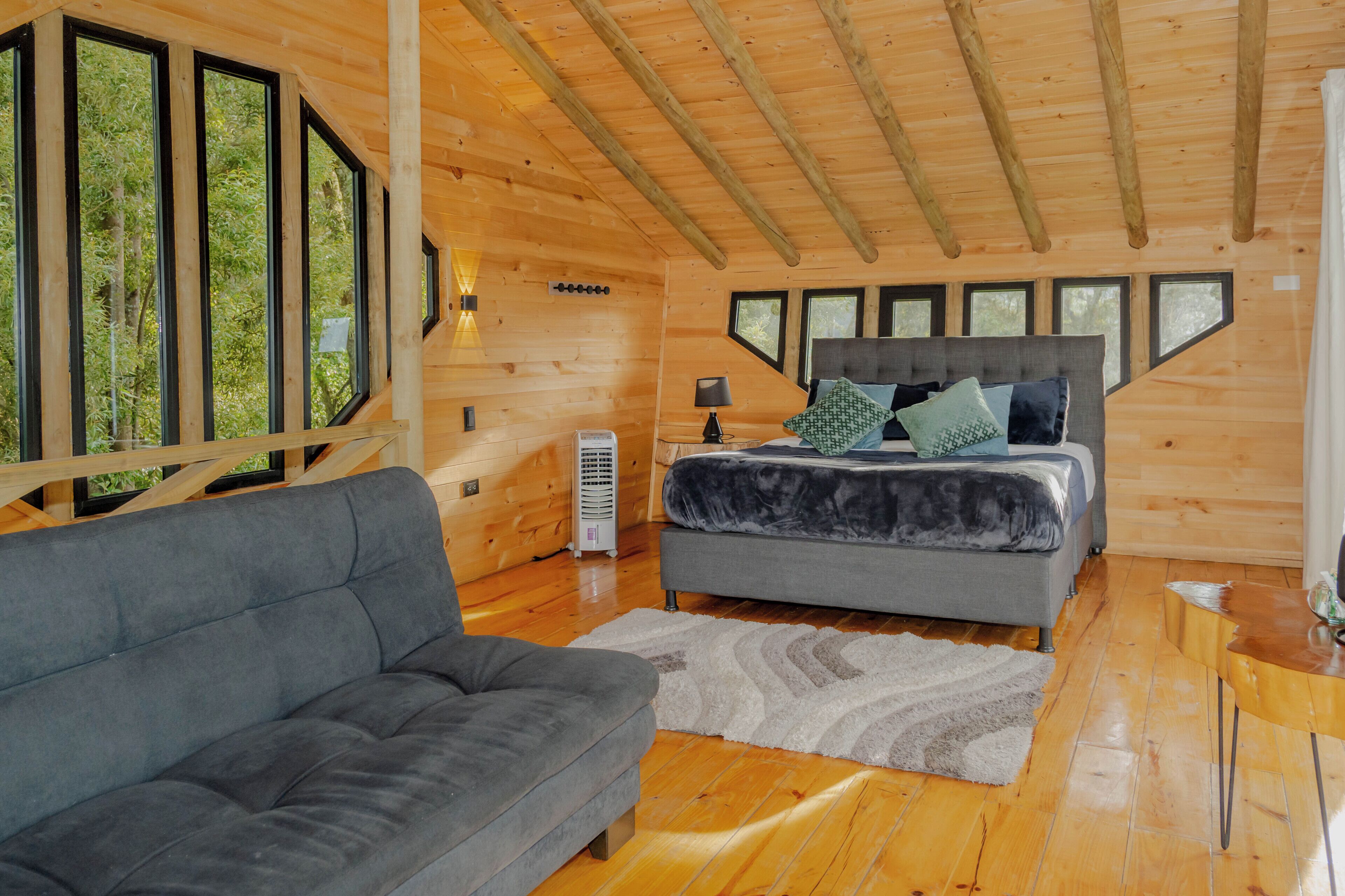 Interior de una cabaña de madera con un sofá, una cama con almohadas, un calentador y ventanas grandes.