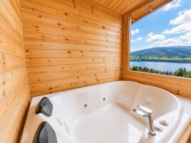 Baño de cabaña de madera con una bañera grande, una ventana que ofrece vistas a un lago y montañas, y techo de madera.