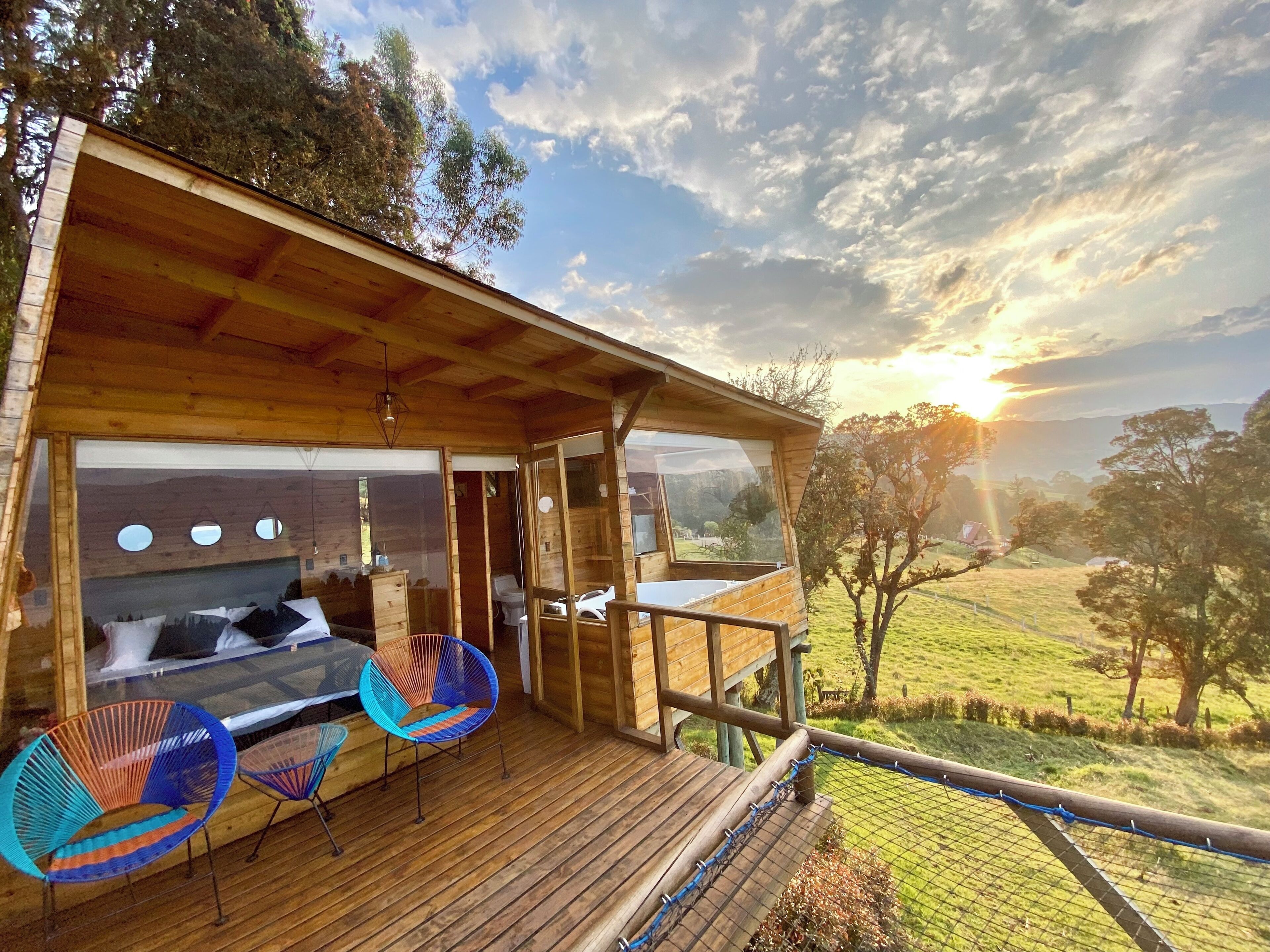 Una cabaña de madera con balcón que da a un paisaje cubierto de hierba, una cama con almohadas y dos sillas coloridas.
