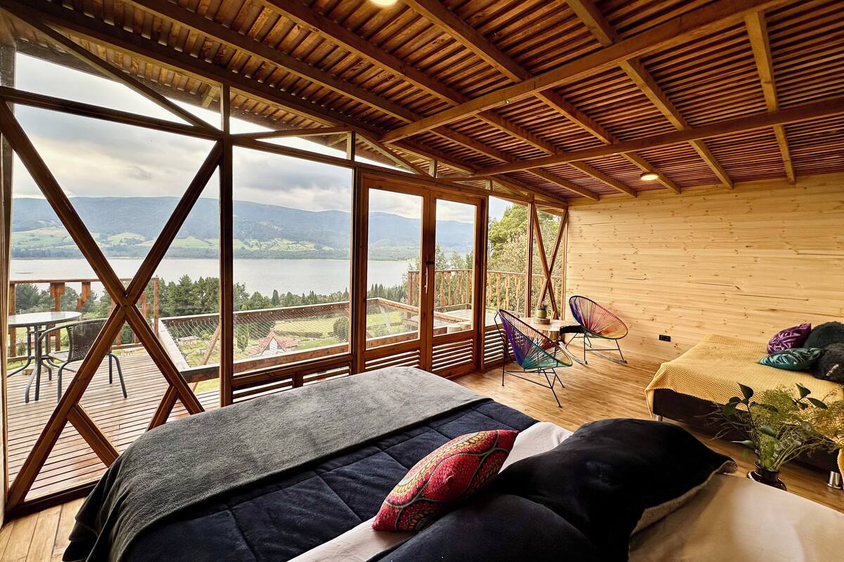 Ein geräumiges Zimmer mit einem großen Bett, zwei Sesseln und Blick auf einen See und Berge durch große Fenster.